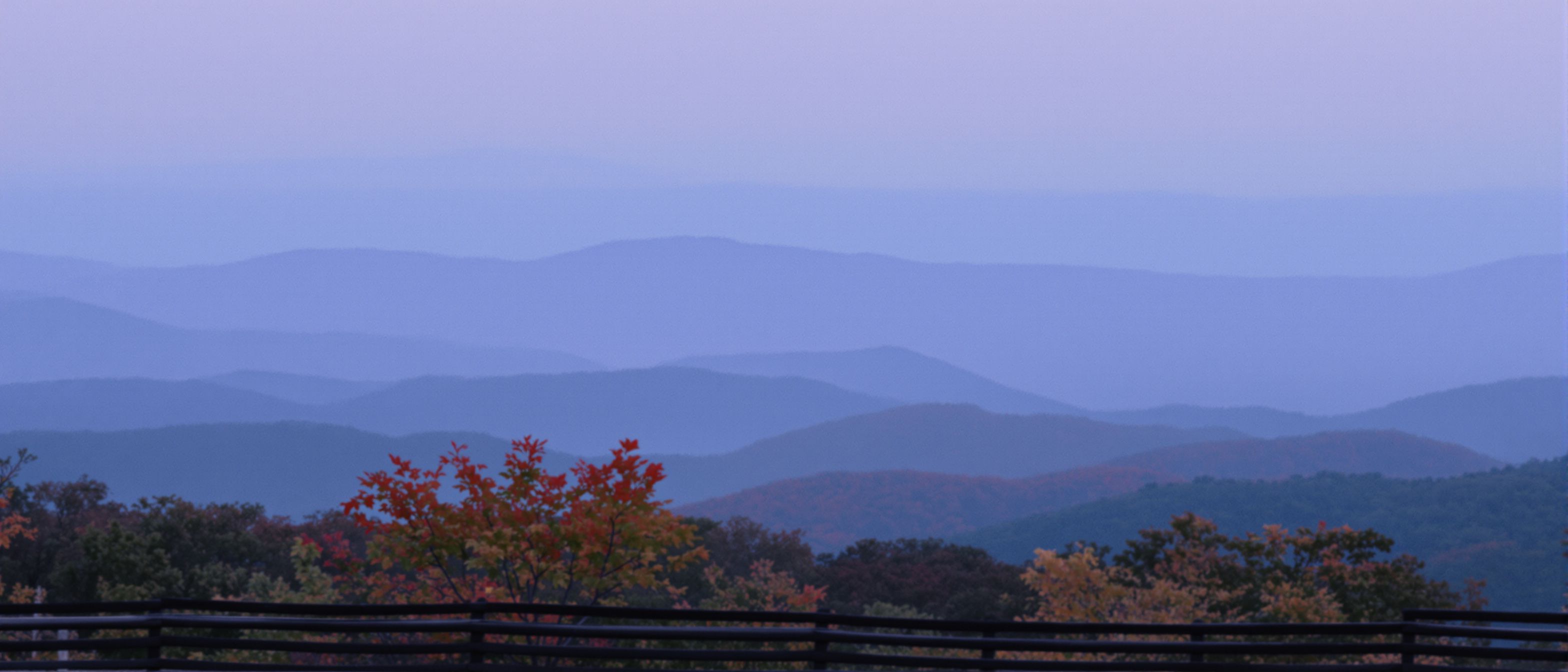 Blue Ridge ridgelines fading into violet haze at dusk, seen over a split-rail fence