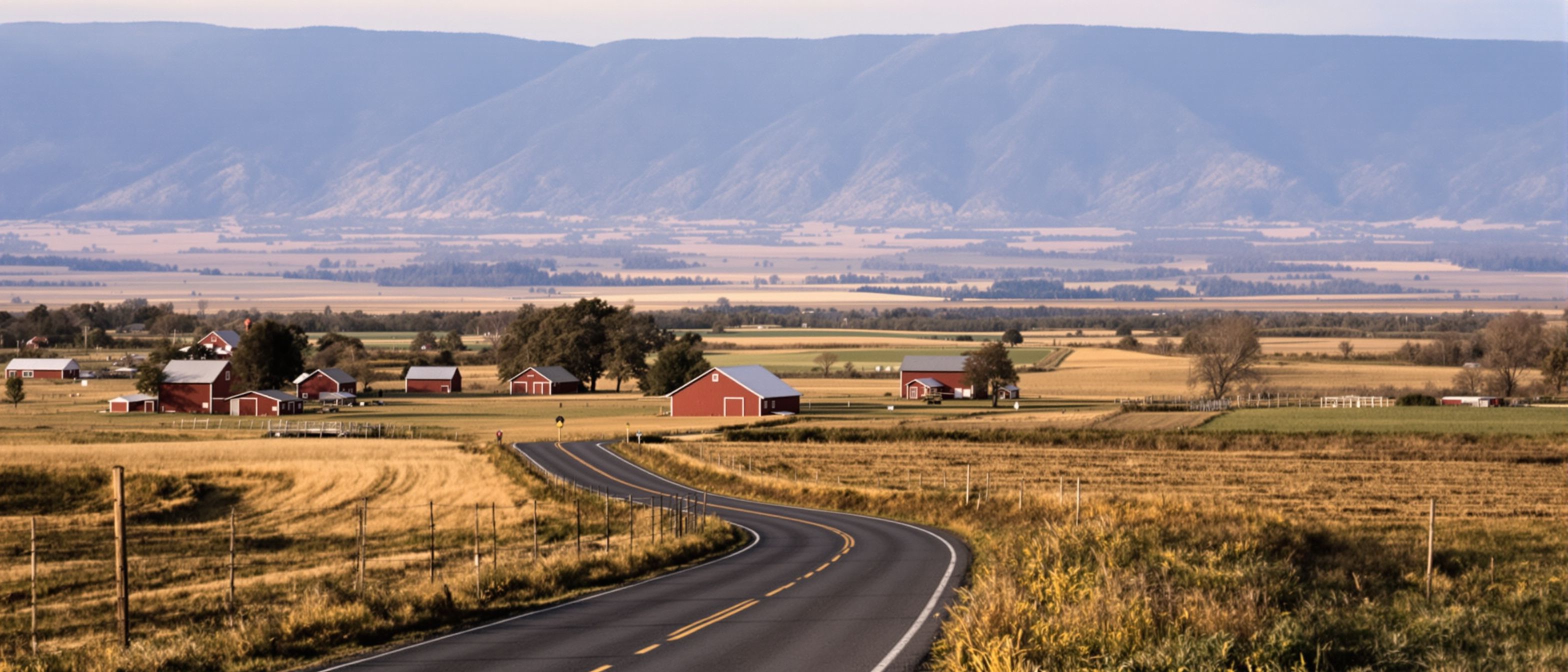 A two-lane road curving past red barns through a broad valley below a mountain ridge